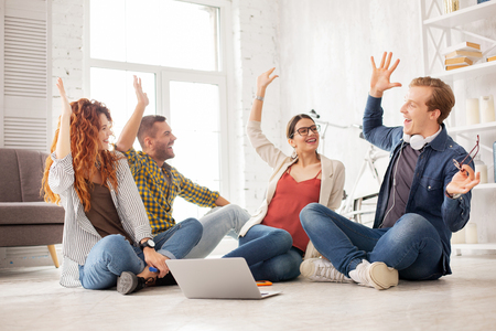 We did it. Cheerful four colleagues rising hands and sitting on floorの写真素材