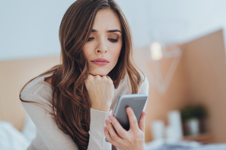 Unpleasant expectation. Sad young woman looking at her smartphone while waiting for a message from her boyfriendの写真素材