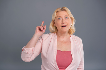 Smart idea. Charming senior woman raising her finger and being excited about having a new idea while standing isolated on a blue-grey backgroundの写真素材