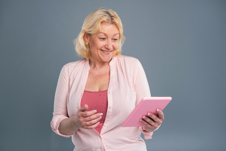 Mood-boosting news. Overjoyed middle-aged woman beaming having read a message on her tablet while standing isolated on a blue-grey backgroundの写真素材
