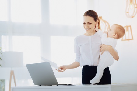 Cheerful woman. Happy young mommy playing with her child and looking at laptopの写真素材