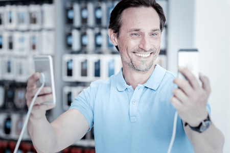 Different models. Delighted joyful smiling man holding two amazing smartphones while choosing between themの写真素材