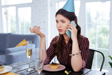 Boyfriend calling. Green-eyed woman wearing festive hat calling her boyfriend while sitting at the table with dessertsの写真素材