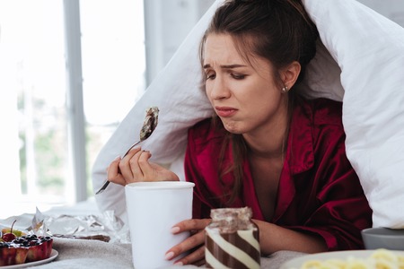 Stressed woman. Stressed crying woman hiding under coverlet while eating ice cream and other desserts after conflict with her man