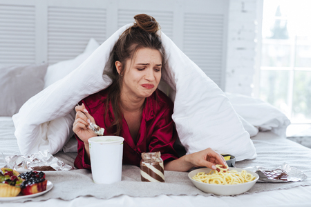 Many snacks. Crying nervous woman lying in her bed surrounding by snacks while feeling awful after breakupの写真素材