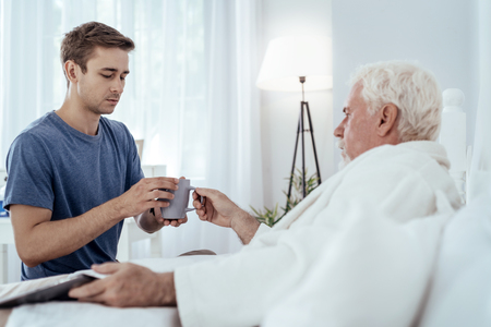 Hot tea. Senior man lying in bed and concentrated man preparing teaの写真素材