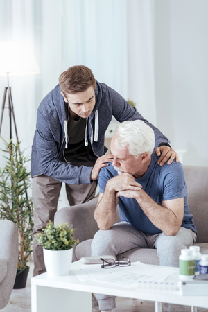 Bad news. Depressed senior man sitting on sofa while man looking at himの写真素材