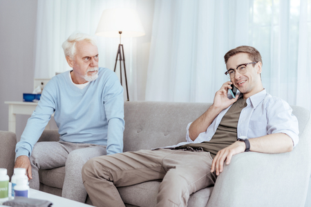 Private life. Cheerful male caregiver using phone while senior man looking at himの写真素材