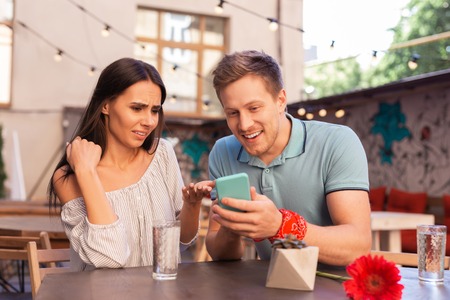 Curious news. Modern good-looking woman and man reading curious news while holding smart phone in...
