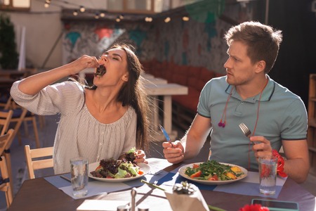Eating lettuce. Dark-haired woman eating some lettuce with her hand while sitting in restaurant along with her boyfriendの写真素材