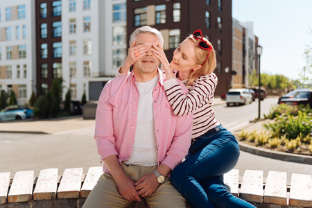 Do not open. Nice joyful woman closing her husbands eyes while preparing a surprise for himの写真素材