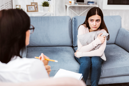 Emotional young woman looking at her therapist and hugging a cushion while feeling worried because of her illnessの写真素材