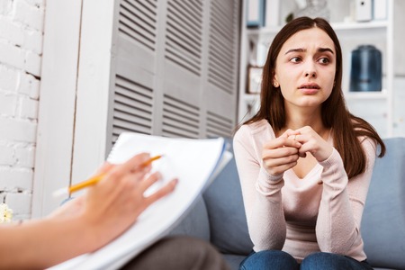 Thoughtful young patient looking into the distance while sitting with her therapist and describing her illnessの写真素材