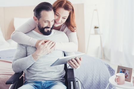Convenient device. Calm optimistic disabled man sitting in a wheelchair with his wife standing behind his back and looking at the modern convenient tabletの写真素材