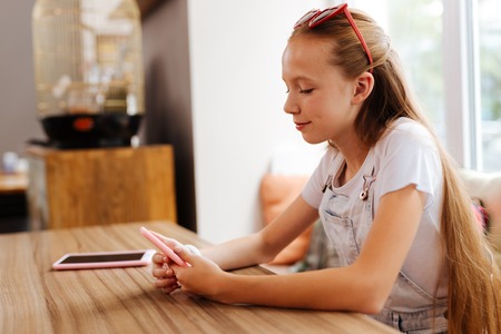 Appealing smiling teenager with blonde long hair reading message on the phone in pink coverの写真素材