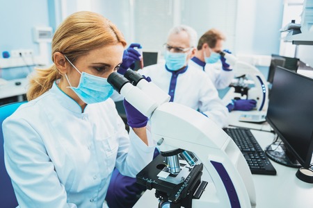 Serious lab assistant sitting in semi position while working with microscopeの写真素材