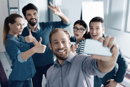 Cheerful bearded young man making selfies and resting with his colleagues while standing in the...