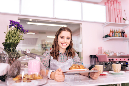 Appealing young barista smiling broadly while preparing two latte and one croissantの写真素材