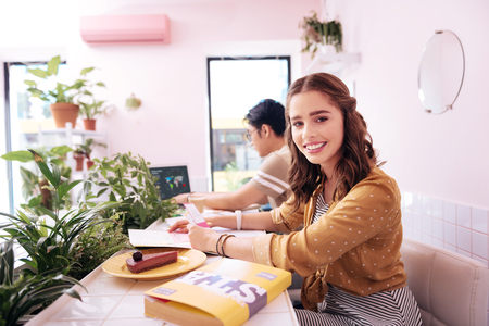 Beaming student wearing stylish striped dress eating cheesecake while sitting in bakeryの写真素材