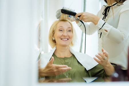 Pleasant skillful woman holding a hairdryer while showing the result to her clientの写真素材