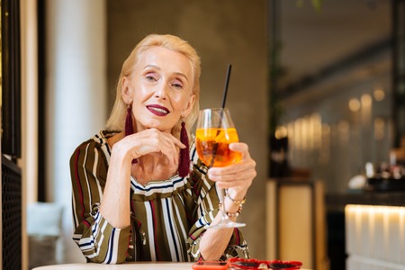 Cheerful nice woman smiling to you while holding a glass of cocktail