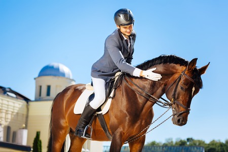 Handsome smiling man wearing helmet and white gloves petting horse while sitting on himの写真素材