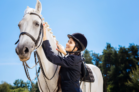 Appealing dark-haired horsewoman smiling broadly while looking at her gentle white horseの写真素材