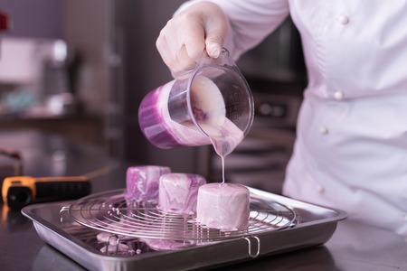 Fruit cakes. Baker wearing white jacket cooking little fruit cakes while pouring some mirror icing on themの写真素材