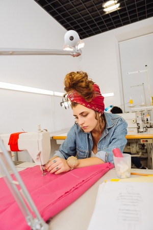Sewing alone. Calm concentrated pretty dressmaker sitting at her workplace in the atelier and sewing...