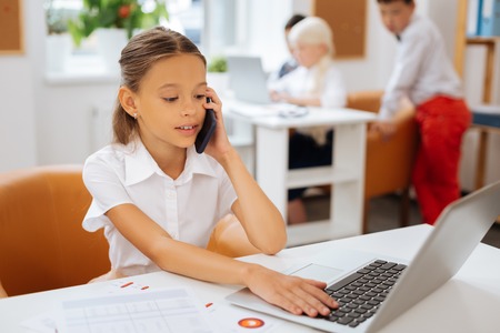 Little manager. Solid pretty schoolgirl speaking on the phone while sitting next to the laptop