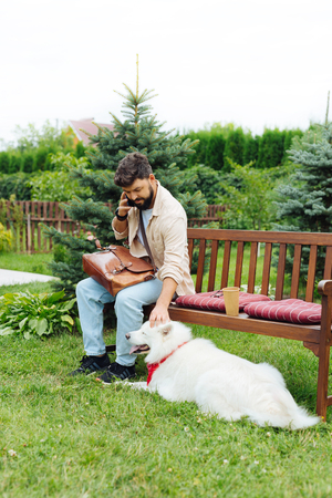 Playing with dog. Dark-haired man playing with his white dog sitting on bench while talking on the phoneの写真素材