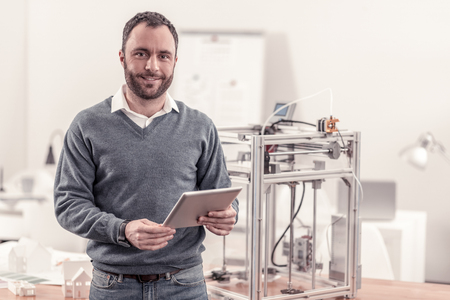 Nice bearded man holding a tablet standing near 3D printerの写真素材