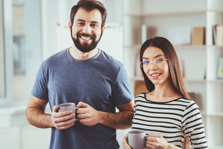 Great mood. Joyful young people looking at you while holing cups of tea in their handsの写真素材