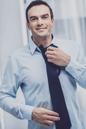 Style. Positive joyful man feeling happy and choosing a tie for a suit before going to work while looking at you with good emotionsの写真素材