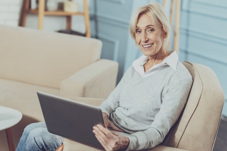 Positive mood. Cheerful aged woman smiling while surfing the Internet