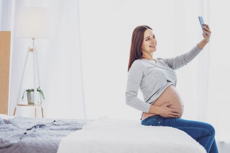 Young mother. Delighted emotional pregnant woman using mobile phone and making selfie while having a...