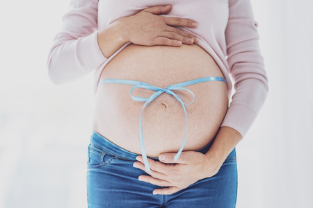 Long waited boy. Close up of young woman with her future child inside standing in front of you and being tied with blue ribbonの写真素材