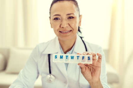 Selective focus on a weekly organizer for medication held by a female doctor smiling cheerfully into the camera.の写真素材