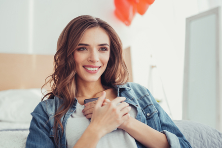 Long waited message. Close up of impressed young girl expressing happiness while sitting in bedroom and keeping a mobile phone at her chestの写真素材