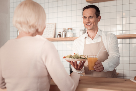 For you. Positive friendly waiter standing at the counter while holding food for cafe customerの写真素材