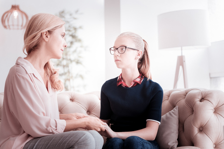 Sincere conversation. Calm serious caring mother sitting on a sofa and looking attentively at her worried daughter while holding her handsの写真素材