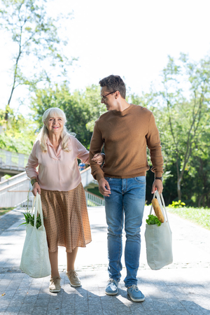 Social care. Joyful brunette man holding shopping bag with products while supporting old womanの写真素材