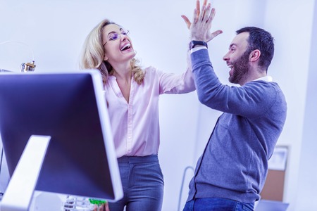 Giving high-five. Cheerful female person standing near workplace and communicating with her colleagueの写真素材