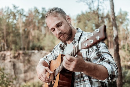 True happiness. Close up of happy handsome bearded man feeling extremely relieved while playing the guitar sitting near mountainsの写真素材
