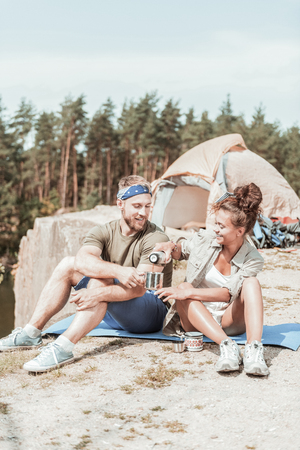 Rest after hiking. Smiling beaming dark-haired woman pouring tea in cup of her boyfriend while having rest after hikingの写真素材
