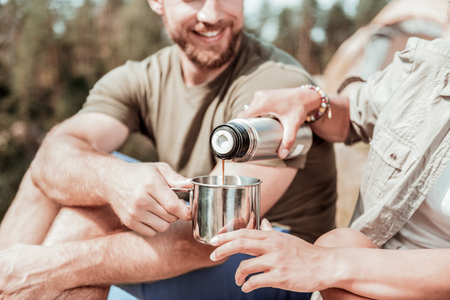 Thermal bottle. Caring girlfriend wearing bracelets holding thermal bottle with tea for her man while having picnicの写真素材