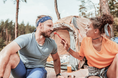 Tasting tomato. Handsome bearded man tasting some fresh tomato while sitting near his appealing girlfriend having picnicの写真素材
