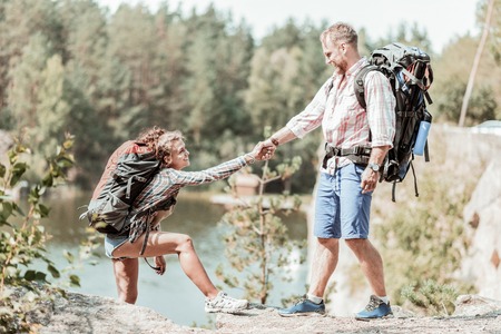 Man helping. Strong bearded man with heavy backpack helping his beaming curly woman reaching the top of rockの写真素材