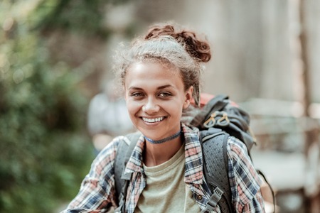 Beaming backpacker. Close up of beaming smiling female backpacker enjoying her day greatly while hiking in mountainsの写真素材