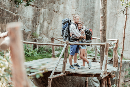 Backpacks and hiking. Caring loving man hugging his appealing tender dark-haired woman while wearing backpacks and hikingの写真素材
